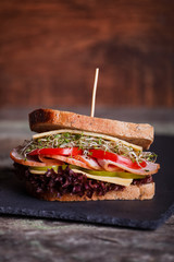 Sandwich with cereals bread with lettuce, slices of fresh tomatoes and sweet pepper, ham, prosciutto, salami, cheese  and  onion sprouts on dark marble background. Close-up