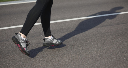 Jogging woman on track. Legs of woman running on road near park or forest. Closeup of female in running shoes going for run on road at sunrise or sunset.