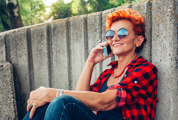Hipster mature woman is calling via cell telephone while sitting in the fresh air in warm spring...
