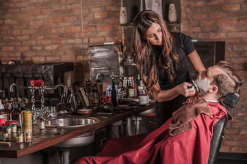 Female barber shaving a client's beard while sitting in a chair in a barber shop