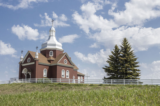 horizontal image of a beautiful brown ukrainian church sitting on a hill with white fence and two spruce trees under a most beautiful blue sky with white clouds floating by in the summer.