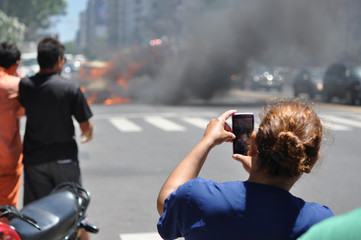 Zuschauerin benutzt Handy um brennendes Auto mitten auf der Straße in Buenos Aires, Argentinien zu filmen - obrazy, fototapety, plakaty