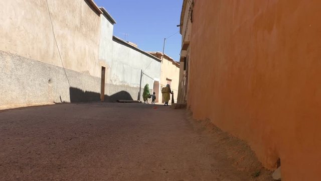 Two local women in traditional muslim clothing and a child walking slowly along quiet Moroccan village street in between pale orange walls on a hot sunny day 