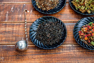 Different kinds of tea on plates on wooden background near vintage strainer. Assortment of dry tea. Tea concept. Tea leaves.  Top view. Closeup