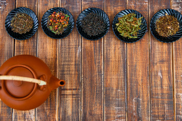 Different kinds of tea on plates near brown teapot on wooden background. Assortment of dry tea. Tea concept. Tea leaves. Frame. Copy space. Top view.