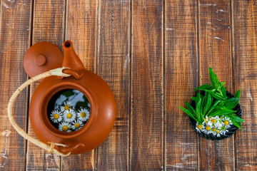 Fototapeta na wymiar Open brown teapot with flower of chamomile near black plate with fresh mint and chamomile on wooden background. Frame. Copy space. Tea concept.