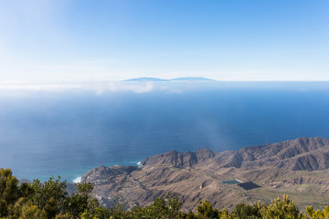Overlook Mirador de Alojera to the canyon Barranco del Mono with the village Alojera. Trade winds with scattered clouds comes from the north into the valley. In the background, the island Las Palmas