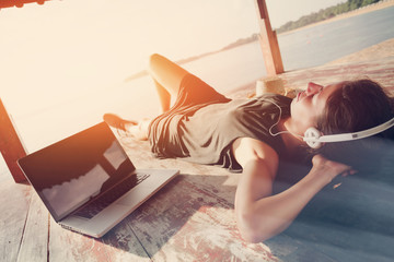 Young woman chilling with laptop and listening music with headphones in shadow shelter. Blurry...