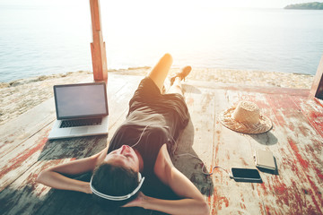 Young woman with laptop resting and listening music with headphones near the ocean. Intentional sun...