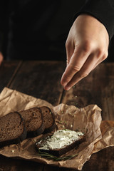 Hand pours some herb spicies on sandwich with rustic bread and soft goat cheese with herbs near slices of homemade luxury rye bread with figs and rosemary spice on craft paper on wooden table