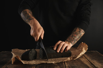 Unrecognizable baker in black sweatshot with tattooed hands cut freshly baked homemade charcoal bread with big chief knife on slices on craft paper on wooden rustic table ready for sale