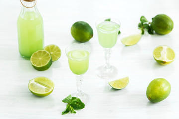 Home lime liquor in a glass and fresh lemons, limes on the white wooden background