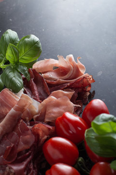Jambon mix. Ham. Traditional Italian and Spanish salting, smoking, dry-cured dish - jamon Serrano and prosciutto crudo sliced with herbs and tomatos on dark stone background. Copy space. Closeup.