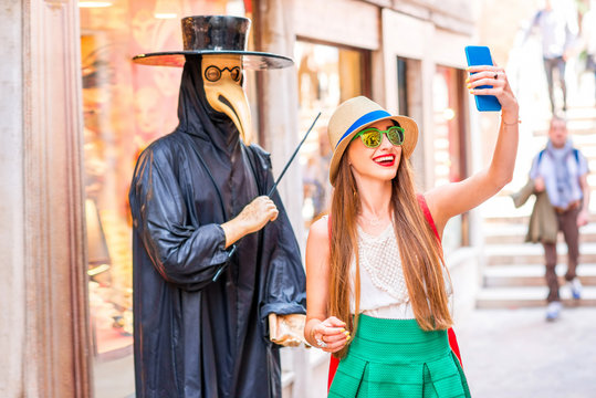 Young female traveler making selfie photo with traditional venice doctor manikin with mask and black costume on the street in Venice