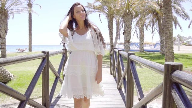Single pretty young Caucasian woman in white dress and long brown hair crossing little bridge at beach with palm trees behind her
