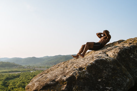 Muscular athletic black man doing press ups relying on the rocky peak. Breathtaking mountain landscape background. Training outdoor concept
