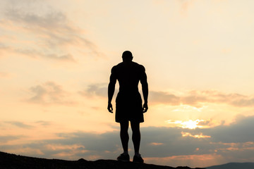 Back view on silhouette of bodybuilder posing at the sunrise or sunset in mountains. Handsome...