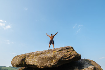 Back view of strong black bodybuilder posing on the rock against blue cloudy sky