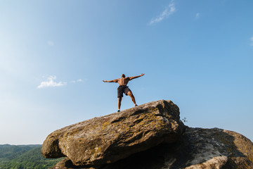 Portrait of strong black bodybuilder with naked torso posing on the rock. Blue cloudy sky background