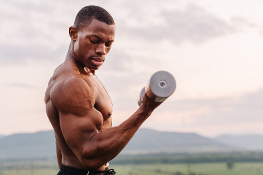 Portrait Of African American Muscular Athlete Lifting Dumbbells Against The Sunset Sky Background