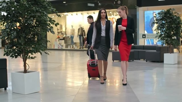 Two beautiful young businesswomen walking across duty-free zone at airport, one of them pulling red suitcase, slow motion shot on Sony NEX 700 