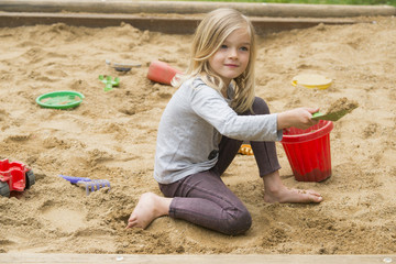 Little blonde girl is playing in sandbox with plastic toy tools