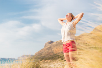 Relaxed woman, arms rised, enjoying sun, freedom and life an a beautiful beach. Young lady feeling...