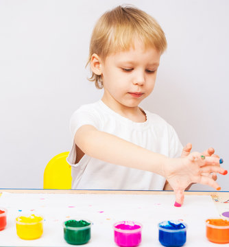 4 years old child painted with hands. Exciting baby boy playing with paint isolated on white background. Boy wearing white t-shirt. Kid sitting at table making colorful hand prints on paper.
