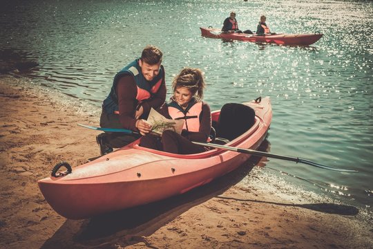 Cheerful friends reading a map in kayaks on a beach.