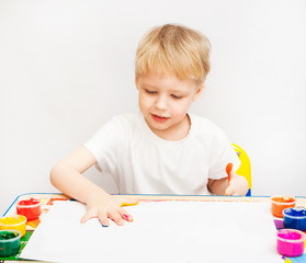 4 years old child painting with hands. Portrait of baby boy playing with paint isolated on white background. Boy wearing white t-shirt. Kid sitting at table making colorful hand prints on blank paper