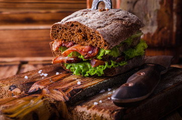Burger sandwich with lettuce, roasted bacon on dark wood cutting board. Selective focus