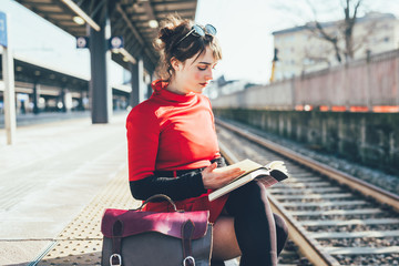 Young beautiful woman sitting on a platform in a train station reading a book- student, commuter, reading concept