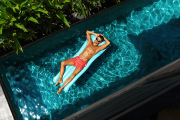 Man On Summer Pool Vacation. Beautiful Handsome Male Model With Sexy Body, Healthy Skin In Swimwear Tanning, Floating On Float Swim Air Mattress In Swimming Pool Water. Recreation, Wellness, Enjoyment