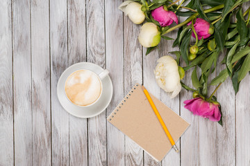 Notebook with a pencil next to coffee and peonies flowers on wooden background. Top view.