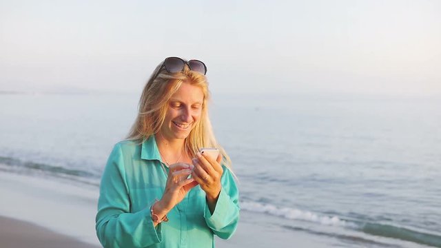 happy  blonde woman smiling texting sms playing app smartphone surfing on social network walking on the beach