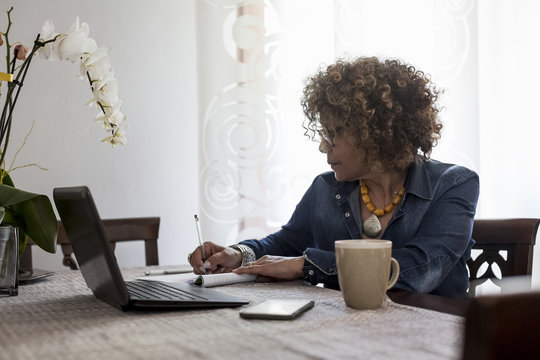 Woman with laptop at home writing in booklet