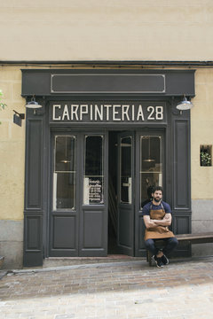 Craftsman with leather apron and arms crossed sitting at the entrance of his workshop