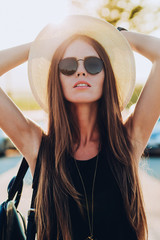 fashion portrait of young brunette woman in a black dress and black sunglasses with a hat on his head against the setting sun