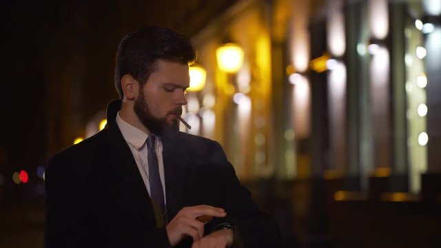 Casual young businessman waiting for someone at night smoking cigarette. 