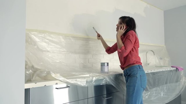 Young woman talking on cellphone and painting wall in kitchen at her new home
