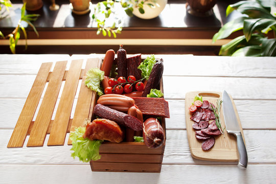 Sausage assortment in wooden box and sliced salami on cutting board with knife. Sausage variety on white wooden background.