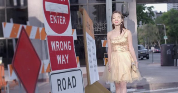 Beautiful young Asian woman wearing a gold sequined dress stops in a patch of sunlight next to road work signs in Downtown Los Angeles. Medium long shot, recorded in slow motion at 60fps.
