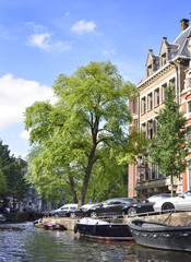 Fototapeta premium Canal of Amsterdam with green trees and boats anchored at the canal. Idyllic water street in Amsterdam, the Netherlands.