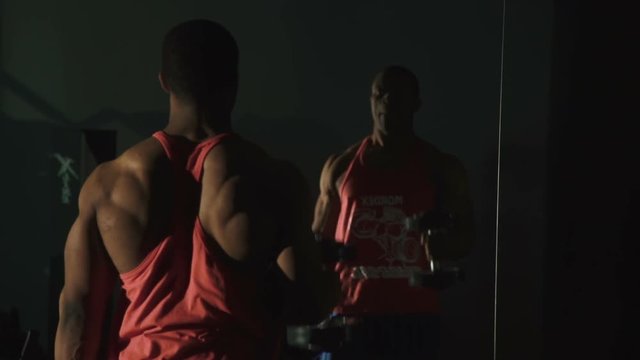 Black muscular african-american weight lifter training at the gym and looks on mirror reflection on dark background 