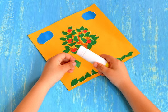 Child makes a torn paper tree. Child holds in his hands a green paper piece and sticks it. Summer tree paper craft. Idea for preschoolers. Home apple tree applique. Step-by-step. Blue wood background.