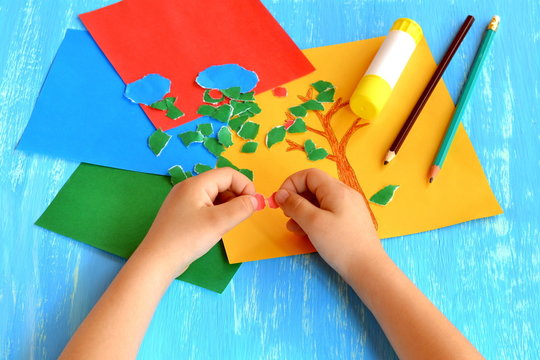 Child tears a red paper into small pieces. Child holds red paper pieces in his hands. Kindergarten art lesson. Set of color paper, pencils, glue stick on wooden background. Fun paper crafts for kids