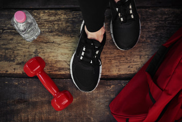 Close-up of woman legs in sportive footwear on wooden floor with dumbbell, bottle and bag. Concept of exercise, fitness and body improvement. 