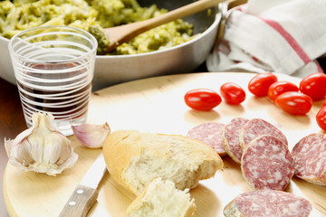 Some slices of salami, bread. tomatoes, knife, garlic and a red wine glass on a wooden cutting board. Background: a pan with broccoli and a cotton rag,
