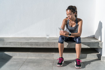 Young woman listening to music with earphones on smart phone app for fitness motivation. Athlete runner in sportswear relaxing sitting getting inspired. Asian mixed race model.