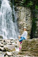 A young girl in a hat sitting on a rock and admires a waterfall in the Carpathian mountains
